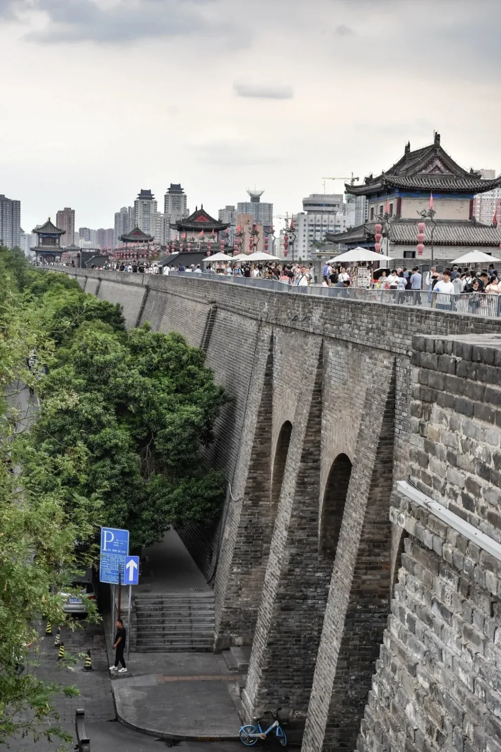 The ancient Xi'an City Wall, made of grey brick with arched buttresses, seen from a street below. On top of the wall, many people walk past traditional Chinese buildings with red roofs and white market umbrellas. Below, green trees line the street where a person stands near a blue parking sign, and a blue bicycle leans against the wall. Modern city buildings are visible in the background.
