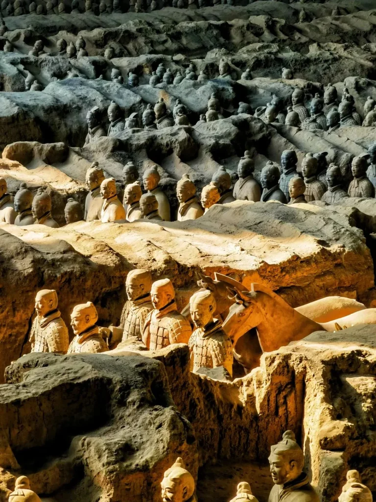 Thousands of ancient terracotta warrior statues and horses stand in formation within a vast archaeological excavation pit. The foreground figures are illuminated by warm light, highlighting their earthy details, while countless others recede into the shaded background.