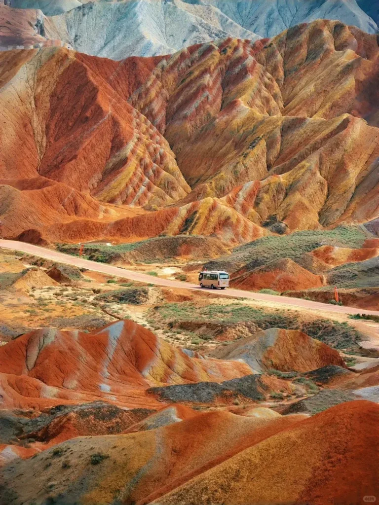 The colorful Zhangye Danxia landform featuring striped rainbow mountains in Gansu, China. The prominent hills show layers of red, orange, yellow, and gray earth. A winding road with a white tourist bus is seen in the middle ground, amidst sparse green and brown vegetation, with distant pale mountains under a light sky.