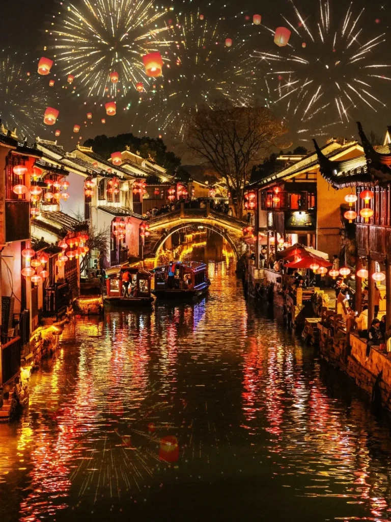A panoramic night view of a traditional Chinese canal town during a celebration. The canal flows through the center, reflecting the warm glow of countless red lanterns decorating the ancient buildings lining its banks. Several traditional boats navigate the water, and an illuminated arched stone bridge connects the two sides. The dark sky is brilliantly lit by bursts of golden fireworks and numerous floating red sky lanterns ascending into the night.