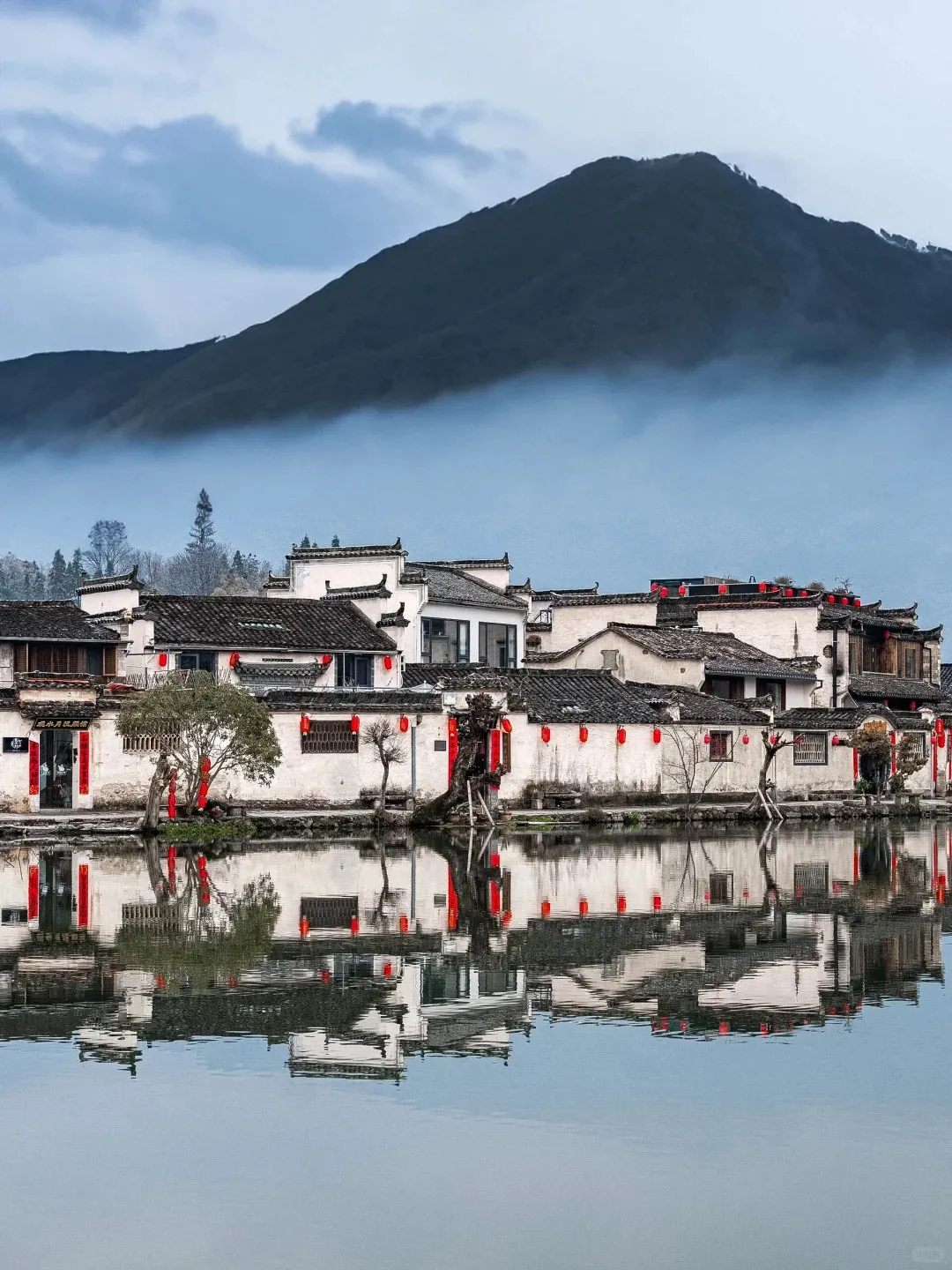 Traditional Huizhou architecture in Hongcun village, Anhui, featuring white walls, black tiled roofs, and vibrant red lanterns, perfectly reflected in a calm body of water. A large, dark mountain is visible in the background, partially obscured by low-hanging mist.