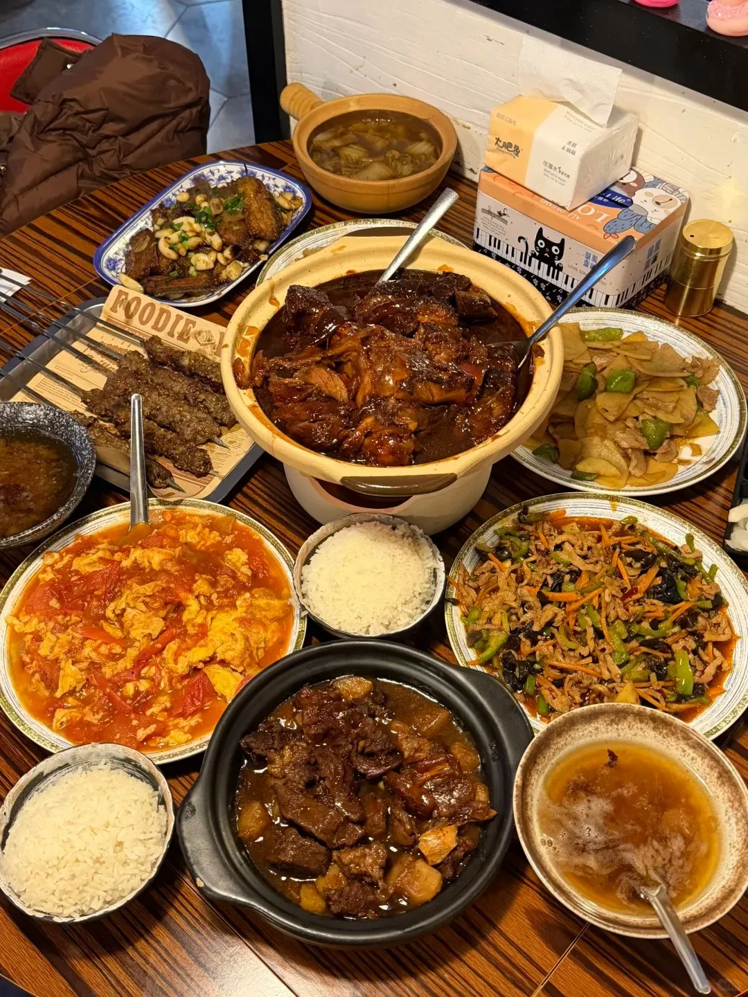 A large wooden table filled with an extensive Chinese meal. Dishes include a large clay pot of dark braised meat, a plate of scrambled eggs with tomatoes, grilled meat skewers, a dish of braised ribs with garlic, a stir-fry of shredded pork with green peppers and wood ear mushrooms, a small wooden pot of vegetable soup, two bowls of white rice, and another smaller pot of braised meat with potatoes.