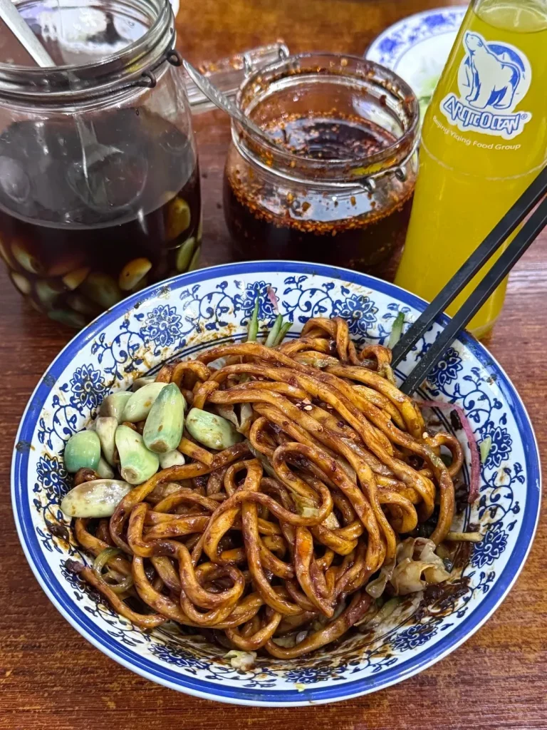 A close-up shot of a bowl of dark sauce noodles (Zhajiangmian) topped with green pickled garlic cloves and shredded vegetables. Accompanying it are a large jar of pickled garlic, a small jar of red chili oil, and a yellow bottle of Arctic Ocean soda, all on a wooden table.