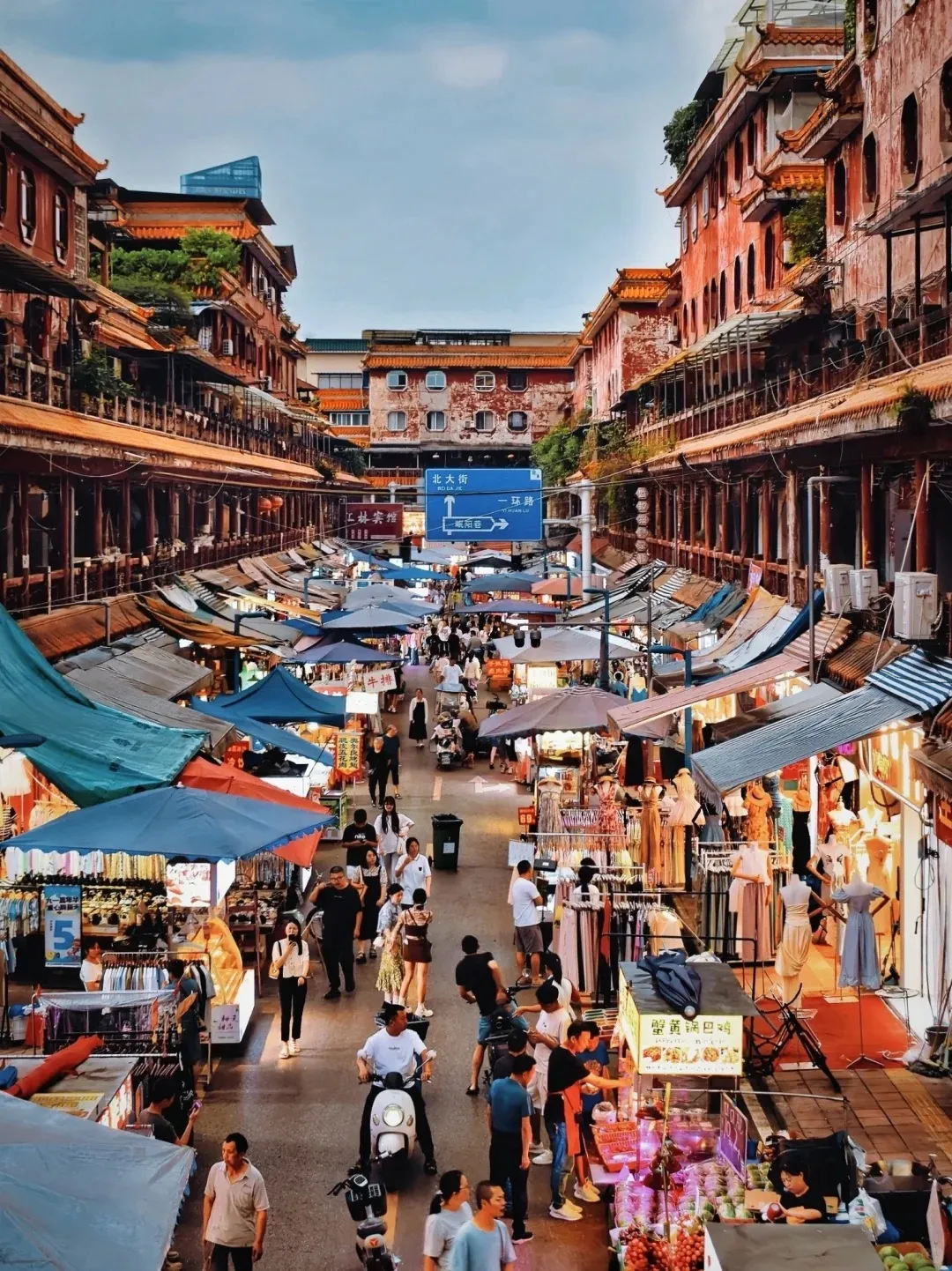 A bustling Chinese market street viewed from above, lined with multi-story traditional red buildings featuring arched windows and wooden balconies. The street below is filled with numerous vendors operating stalls under colorful awnings, selling various goods like clothes and food. Many people walk, shop, and socialize along the crowded street, with a white scooter visible in the foreground. Blue street signs with Chinese characters are mounted on a pole in the middle distance.