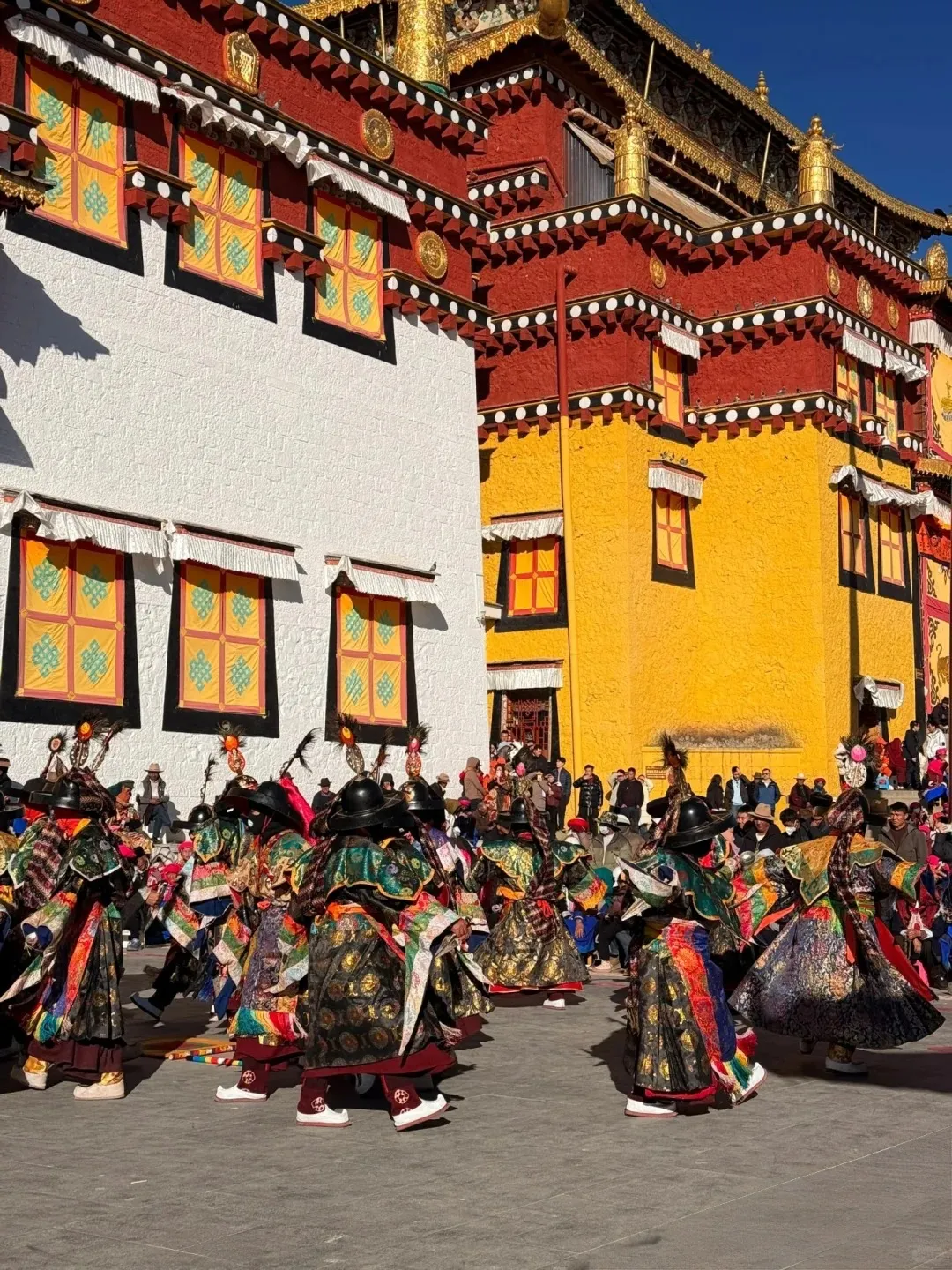 A group of elaborately dressed dancers in colorful traditional robes and black hats perform a ritual dance on a paved outdoor courtyard. Behind them are large, multi-story monastery buildings with vibrant red, white, and yellow walls, ornate windows, and gilded golden rooftops against a clear blue sky.