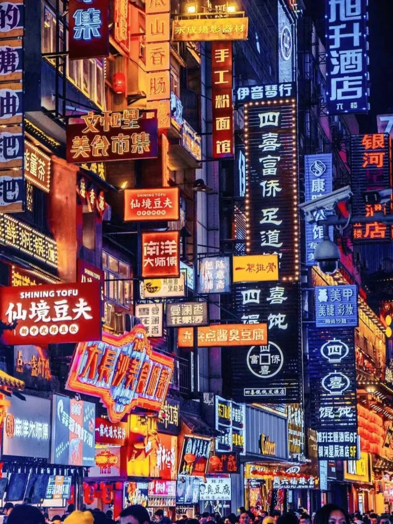 A vibrant night scene in a bustling city street lined with tall buildings adorned with numerous glowing neon and illuminated signs in Chinese characters. Many signs advertise food, including 'SHINING TOFU' (仙境豆夫), 'Stinky Tofu' (臭豆腐), 'Food Market' (美食市集), and noodle shops. A dense crowd of people fills the street at the bottom, enjoying the bright, colorful lights.