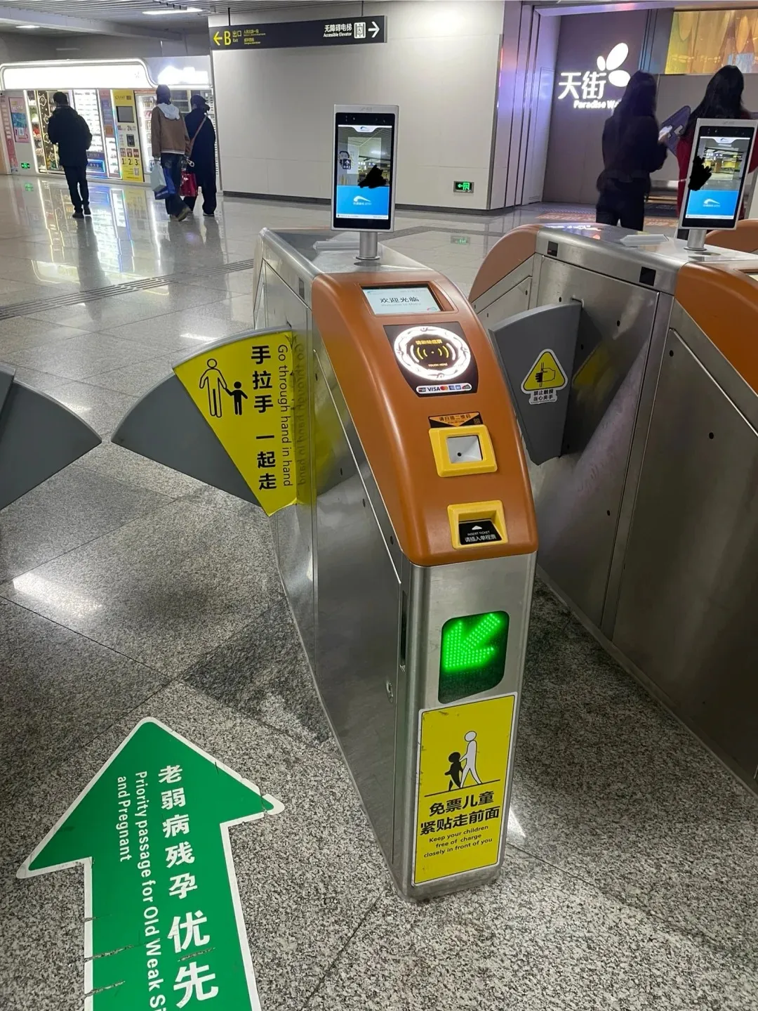 A modern Chengdu Metro station entry point with metal and orange turnstiles. The central turnstile features a screen with 'Welcome', a circular scanner for QR codes, and a tablet for face recognition. A green arrow indicates an open passage. Green floor signage marks a 'Priority passage for Old Weak Sick Disabled and Pregnant', while yellow signs on the turnstiles offer instructions for accompanying children and hand-in-hand passage.