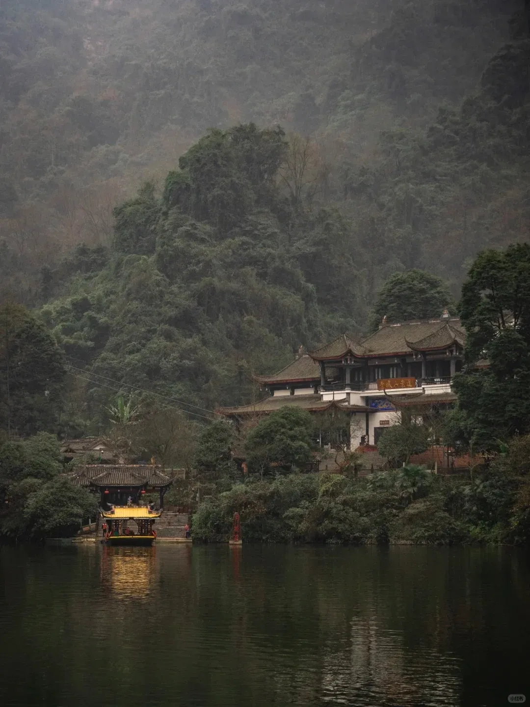 A traditional multi-story Taoist temple with dark tiled roofs stands by a calm lake. A smaller golden-roofed pavilion or boat is docked close to the water. In the background, lush, misty green mountains covered in dense forest rise.