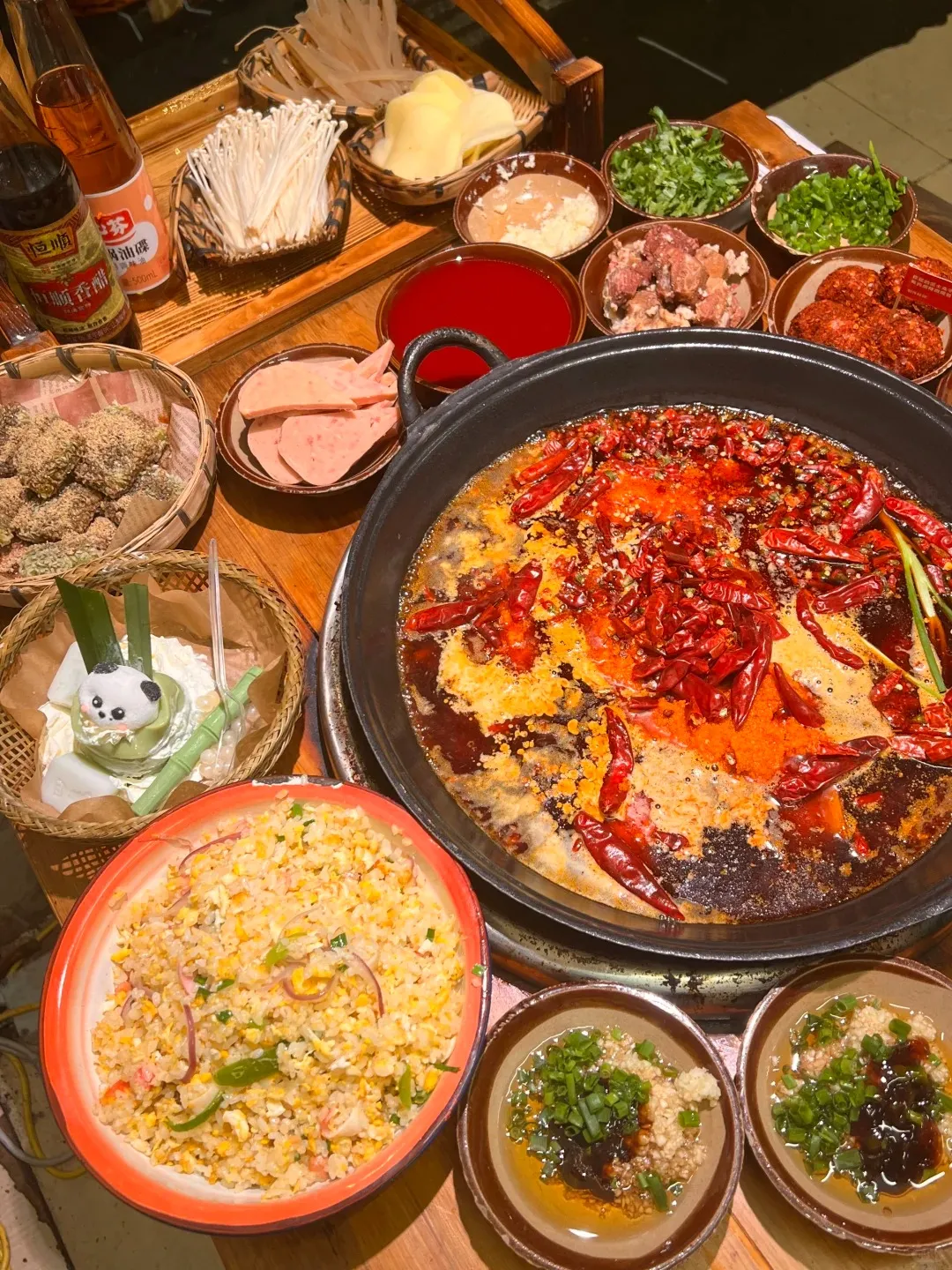 A large black hotpot sits on a wooden table, filled with bubbling red Sichuan chili broth abundant with dried red chili peppers and oil. Surrounding the hotpot are numerous small bowls containing raw ingredients such as thinly sliced meat, enoki mushrooms, wide transparent noodles, sliced ham, chopped cilantro, and chopped green onions. Other dishes include a basket of fried sesame-crusted balls, a bowl of fried rice with egg, corn, and red onions, a small white dessert with a panda face, and dipping sauces with garlic and scallions.