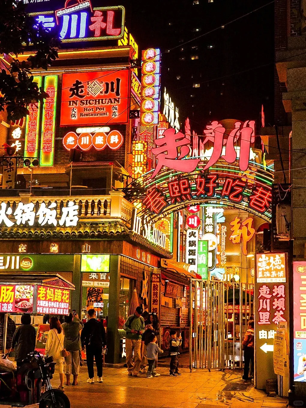 A bustling night street in Chengdu, China, illuminated by colorful neon signs in red, blue, green, and pink. Prominent signs advertise 'IN SICHUAN Hot Pot Restaurant' and other Chinese characters for hot pot and food. People are walking along the wet pavement past various shops and food stalls.