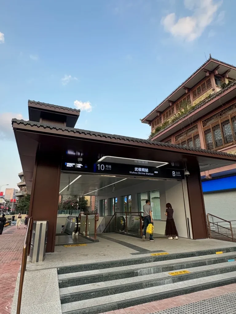 A modern brown metro station entrance, labeled 'Wuhou Shrine Station Line 10' in Chinese and English, with visible escalators and stairs. People are walking near the entrance. Traditional Chinese-style buildings with intricate roofs are visible in the background under a blue sky.