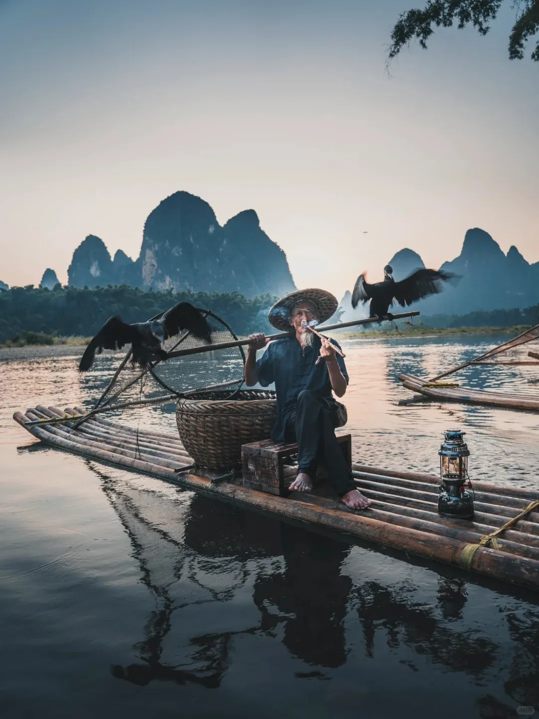 An elderly Chinese cormorant fisherman in a straw hat smokes a pipe while sitting on a bamboo raft. Two dark cormorants are perched on a long pole he holds, one on the left and one on the right with its wings spread. Dramatic karst mountains rise in the misty background, reflecting in the calm river water under a soft, golden sky. A woven basket and a lit lantern are on the raft.