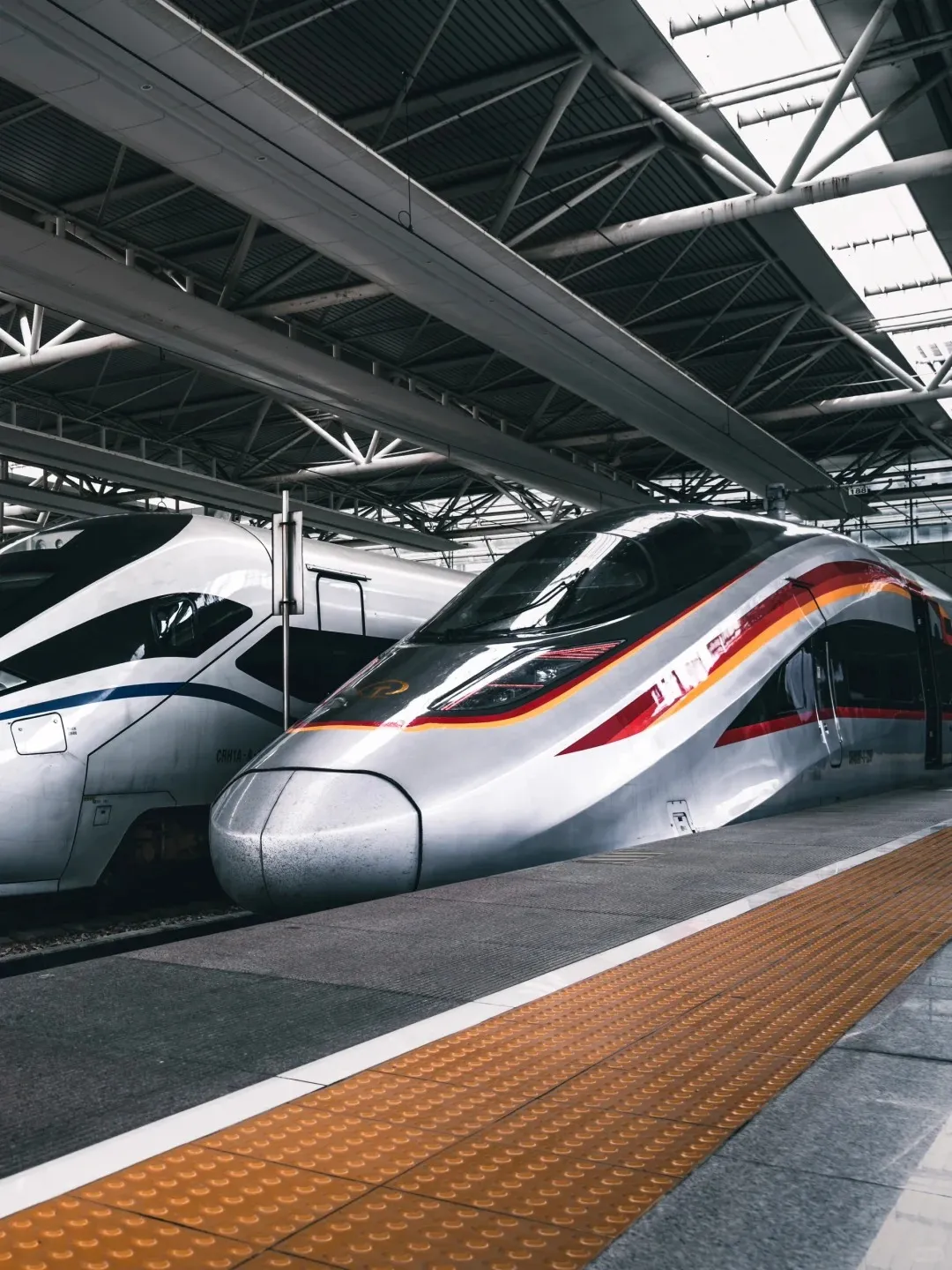 Two modern high-speed trains are parked side-by-side at a train station platform. The train on the right is silver with red and orange stripes, and the train on the left is white with blue stripes. A yellow tactile paving strip is visible on the platform in the foreground, with the station's structured roof overhead.