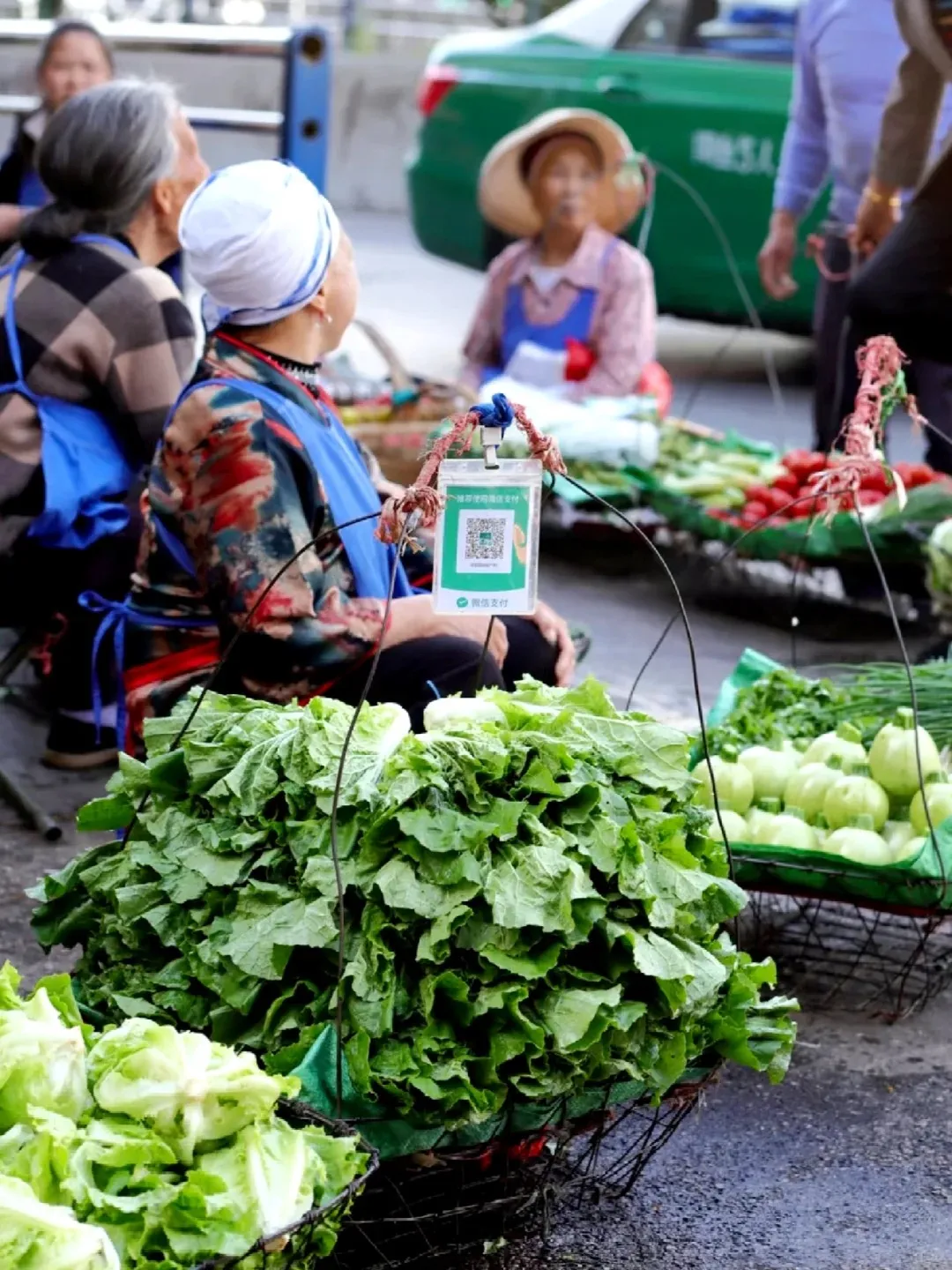 An elderly female vendor sits beside baskets of fresh green leafy vegetables and light green squash at an outdoor market in China, holding up a green card with a WeChat Pay QR code. Other market-goers and a green taxi are visible in the soft-focus background.
