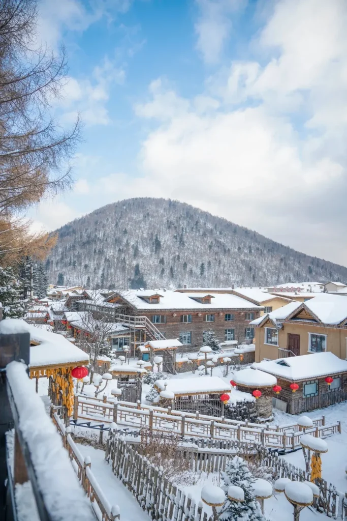 An aerial view of China Snow Town in winter, showing traditional wooden houses with thick, rounded snow on their roofs, fences, and decorative elements. Red lanterns and hanging corn cobs adorn some buildings. A large, snow-covered forested mountain rises in the background under a blue sky with white clouds.