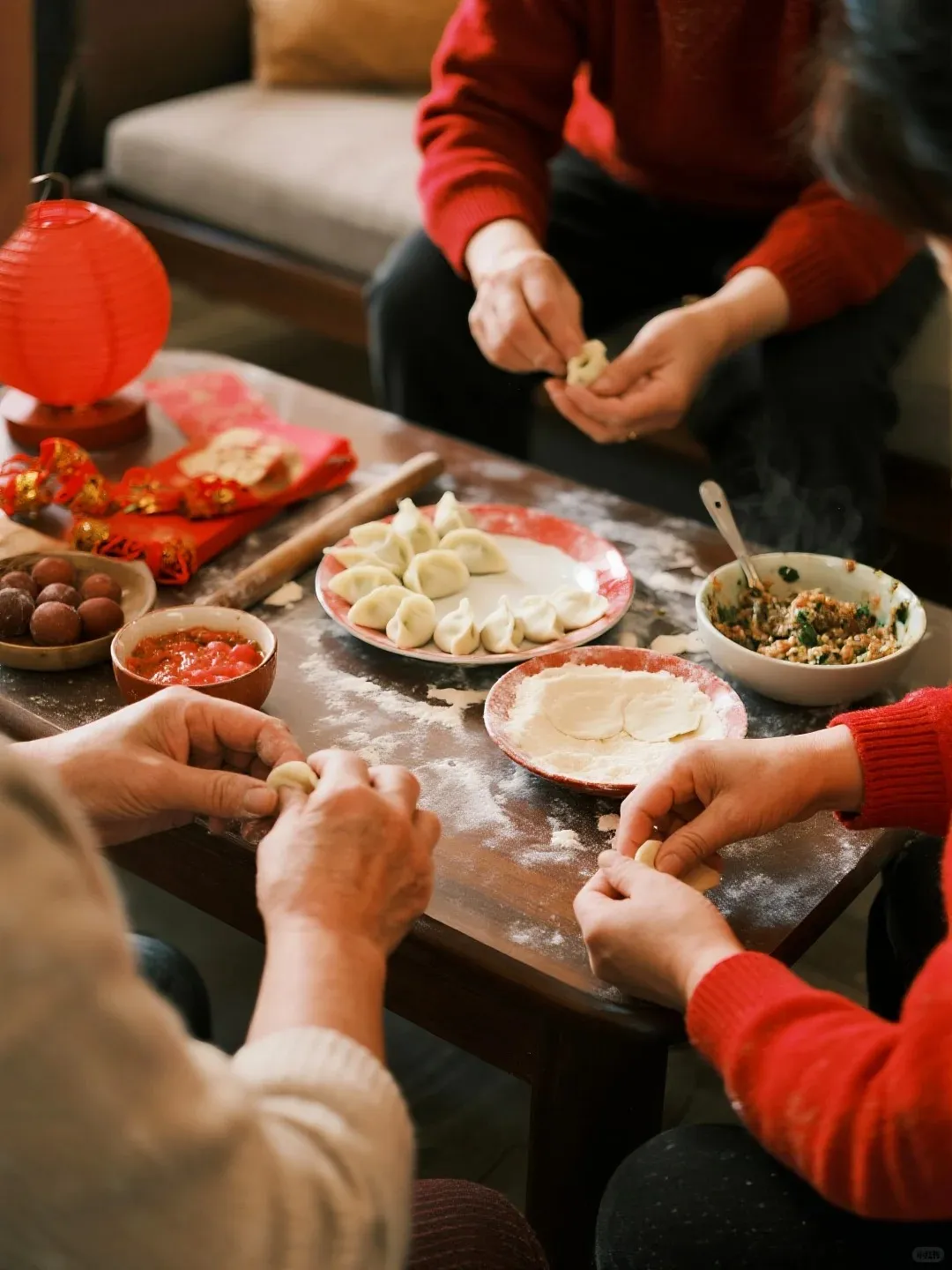 Close-up view of multiple hands from a family gathered around a wooden table, actively shaping Chinese dumplings. The table is dusted with flour and displays plates of completed dumplings, a bowl of savory dumpling filling, a plate of round dumpling wrappers, a wooden rolling pin, a red paper lantern, red celebratory envelopes, a bowl of red sauce, and a bowl of small red, round snacks.