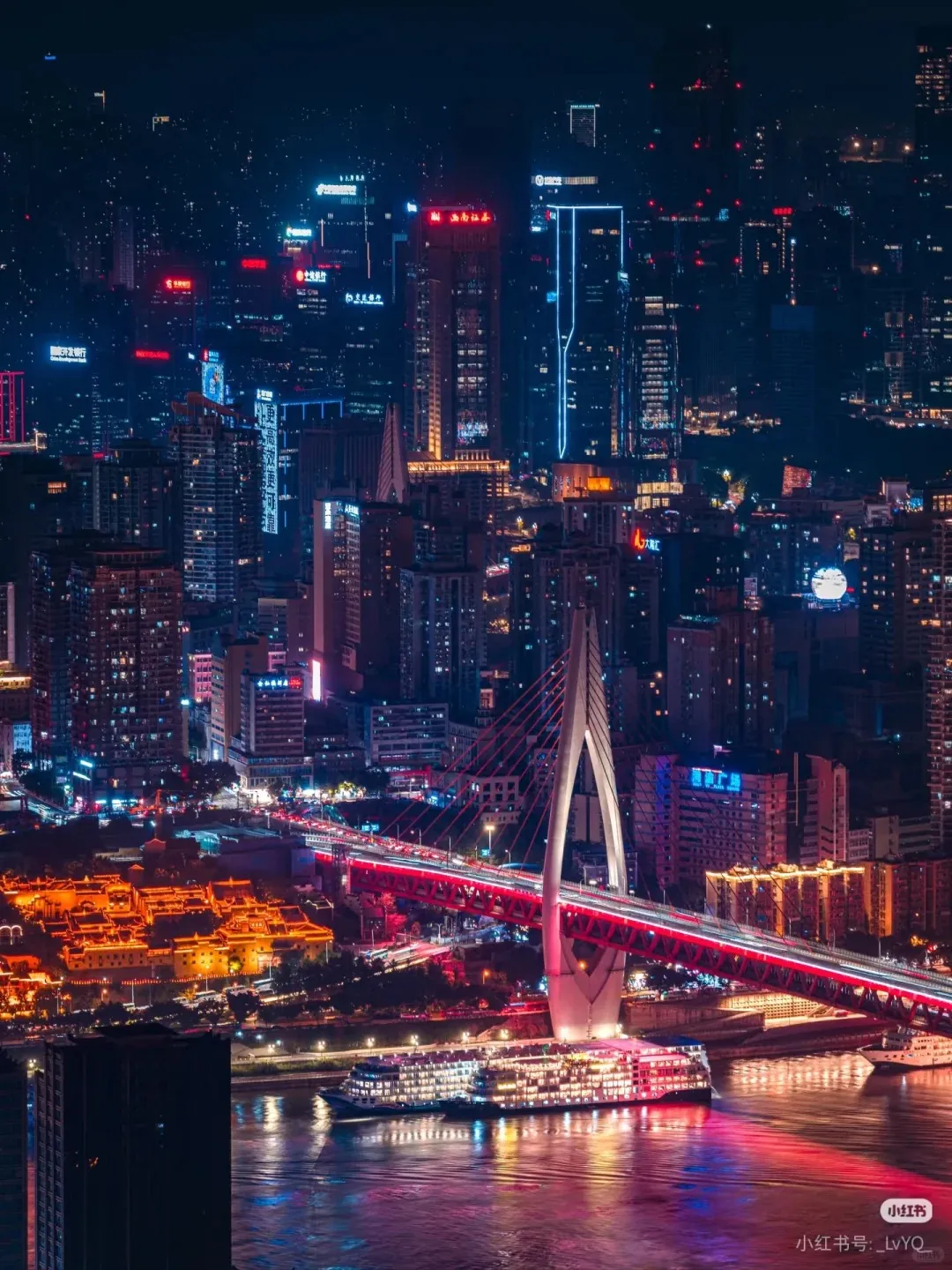 A vibrant night cityscape of Chongqing, China, featuring a large red cable-stayed bridge spanning a river with illuminated cruise ships. The background is filled with numerous skyscrapers and buildings glowing with red, blue, and orange neon lights, along with a complex of traditional buildings lit in warm orange on the riverbank.