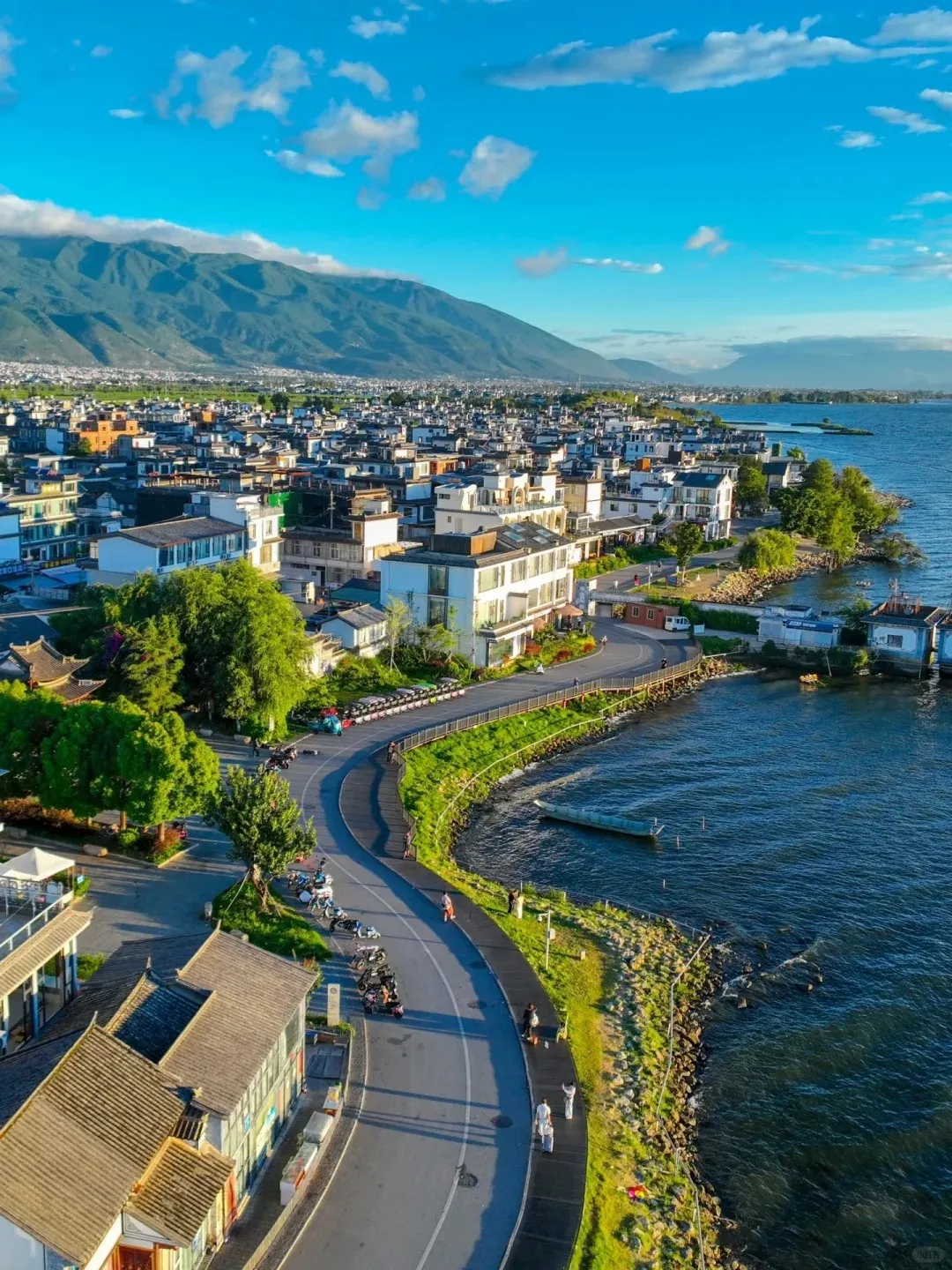 Aerial view of a town with traditional buildings along a winding road and pedestrian path next to Erhai Lake in Dali, Yunnan. The Cangshan mountains are in the background under a blue sky with white clouds. People are walking on the path and motorcycles are parked.