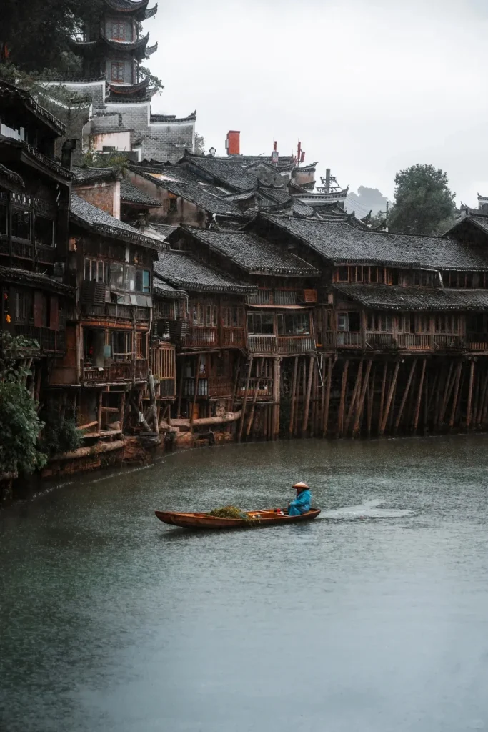 A person in a conical hat and blue poncho rows a wooden boat on the Tuojiang River in Fenghuang Ancient Town. Along the riverbank, traditional dark wooden stilted houses stand, with a pagoda visible on a distant hill under a rainy, overcast sky.