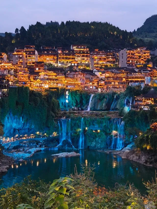 Night view of Furong Ancient Town (芙蓉镇) in China, showcasing traditional multi-story wooden buildings illuminated with warm golden lights built into a cliffside. Below the town, a multi-tiered waterfall, lit with vibrant blue and teal lights, cascades into a dark river that reflects the glow. Lush green foliage covers the surrounding landscape and distant hills under a twilight sky.