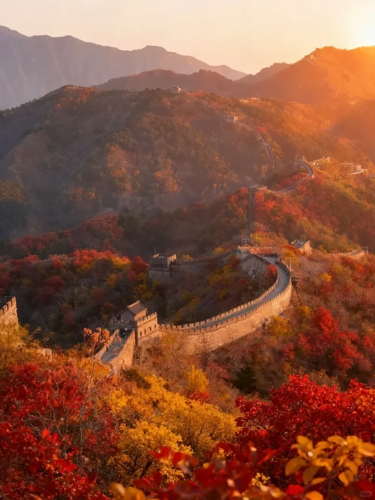 The Great Wall of China winds along a mountainous ridge covered in vibrant red and yellow autumn trees, illuminated by the warm light of a sunset. Several stone watchtowers are visible along the wall, which stretches into the hazy distance.