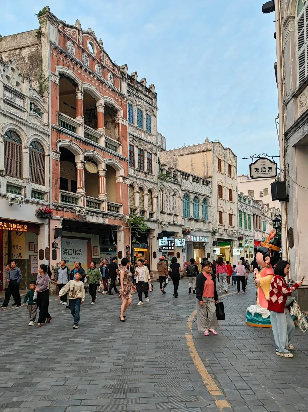 A bustling pedestrian street in Haikou Qilou Old Street, Hainan, lined with historic multi-story buildings featuring arched balconies, elaborate facades, and colonial-era architecture. Many people are walking along the paved street. A sign on the right reads '天后宫 / Tianhou Temple'.
