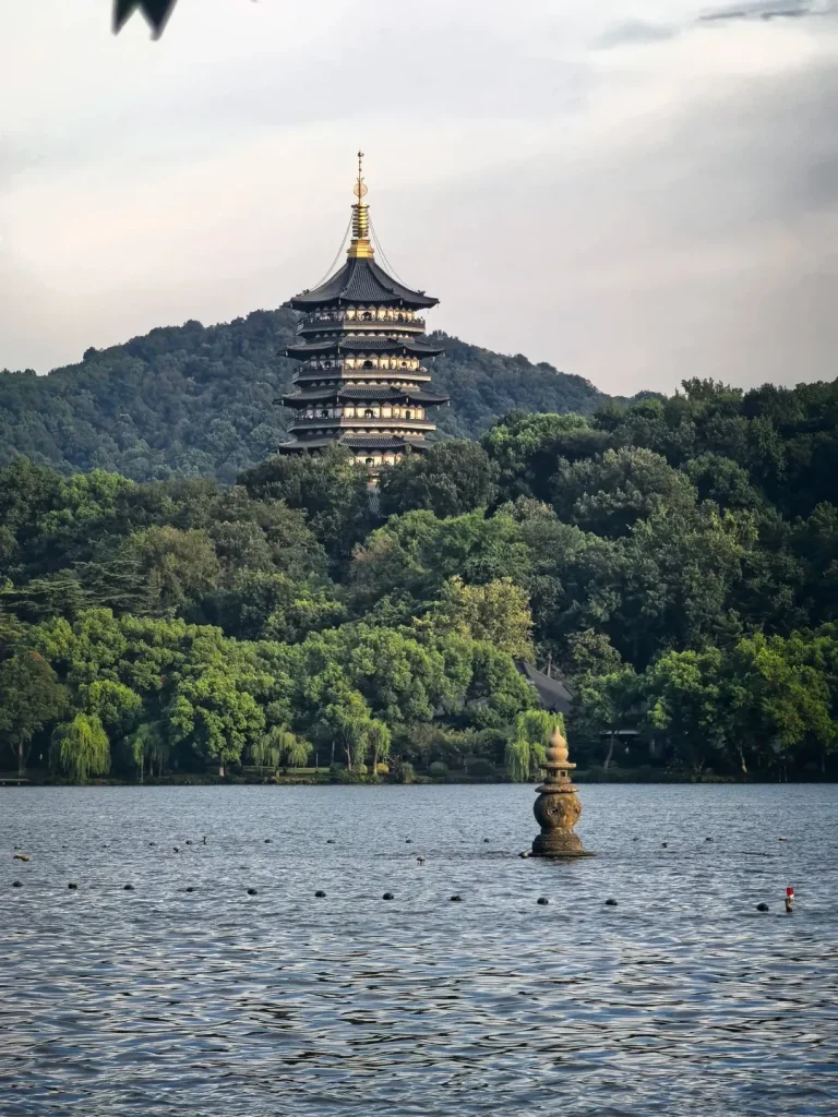 Multi-tiered Leifeng Pagoda with a golden spire, standing on a lush green hill overlooking the calm waters of West Lake. A stone lantern is partially submerged in the lake's foreground, under an overcast sky.