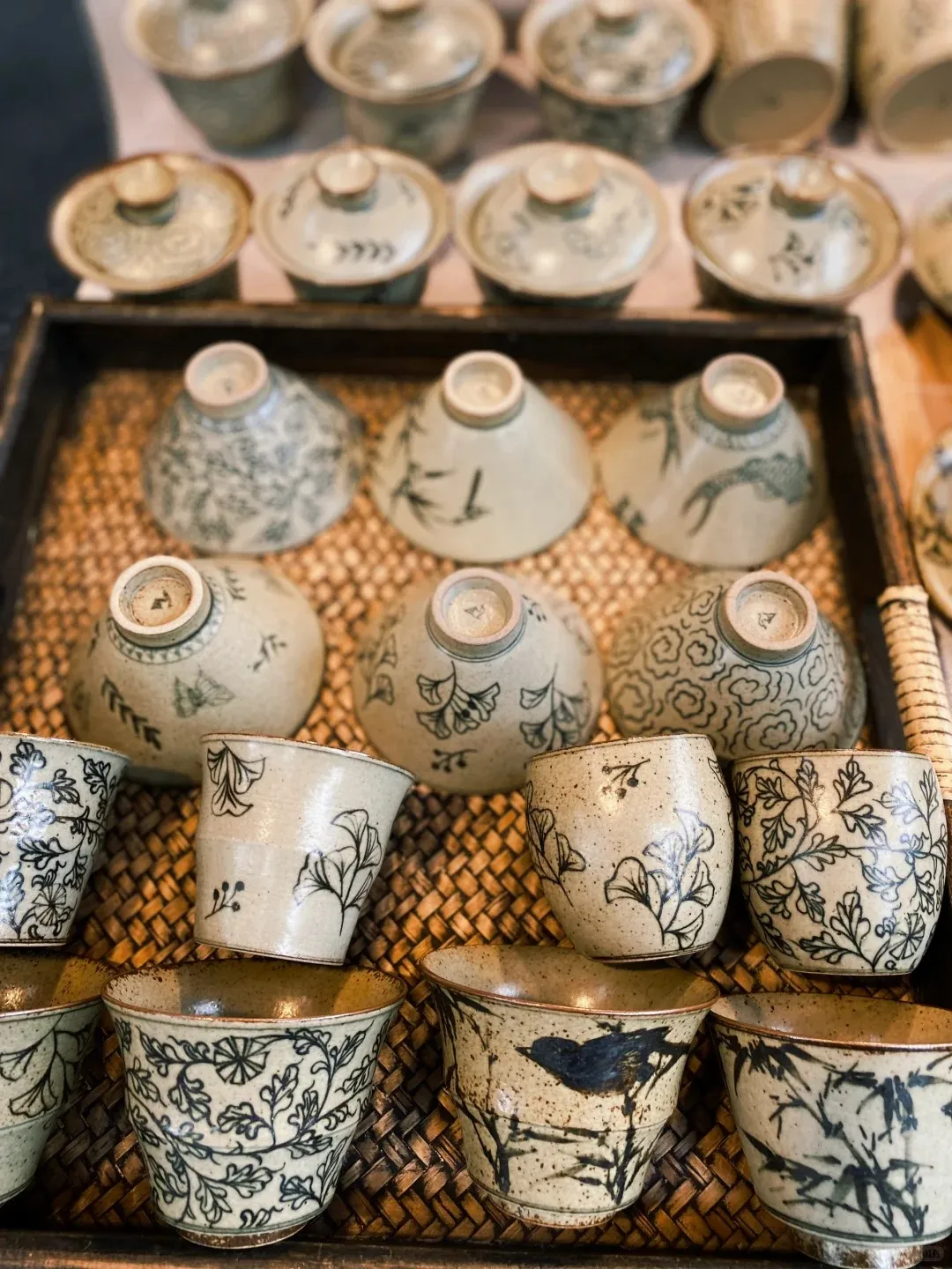 A collection of handmade ceramic bowls and cups with hand-painted patterns, displayed on a woven mat in a dark wooden tray. The pottery is off-white or pale green, featuring black designs of leaves, flowers, bamboo, and a bird. Some pieces are lidded, others are open cups, and several bowls are inverted.