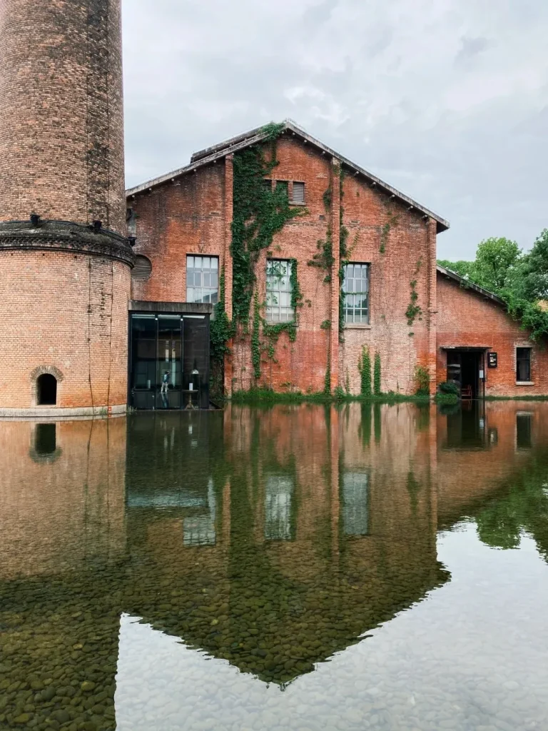 A large red brick building with a tall brick chimney, partially covered in green ivy, reflected in clear water with visible pebbles at the bottom.