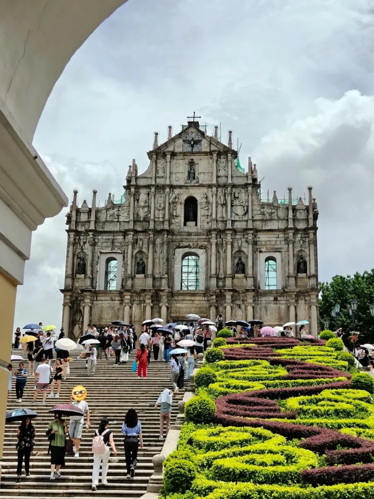 The historical stone facade of the Ruins of St. Paul's in Macau, featuring intricate carvings and statuary, stands at the top of a grand, wide staircase. Numerous tourists are visible on the steps, some holding umbrellas, and sculpted green and reddish-purple hedges line the right side of the staircase. A white architectural arch partially frames the view from the upper left.