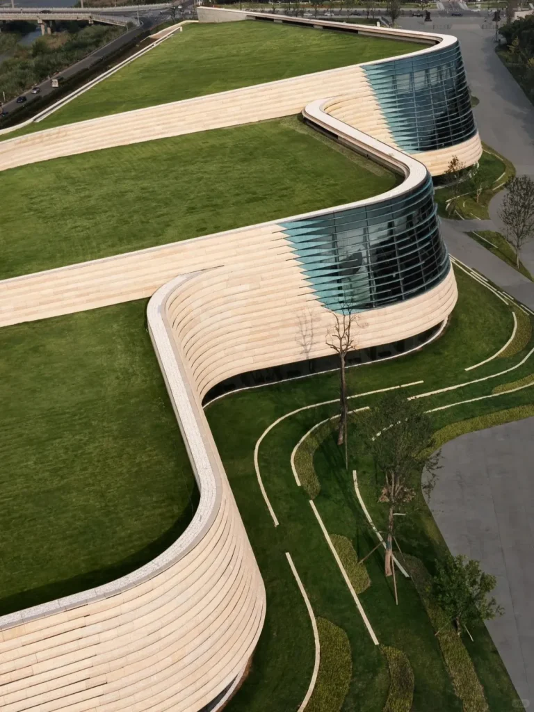 Aerial view of the new Sanxingdui Museum building, featuring two prominent curved structures with extensive green grass roofs and light-colored, layered stone facades. Large, floor-to-ceiling glass sections reflect the surroundings. The landscaped grounds include winding paths and trees, with a river and bridge visible in the background.