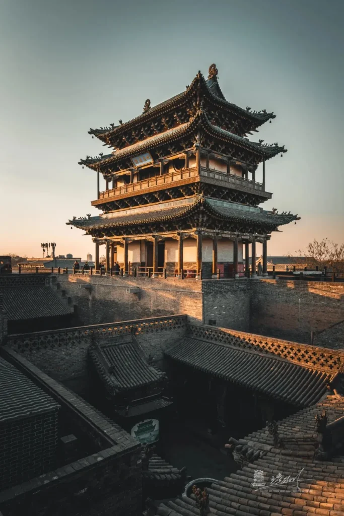 A grand, multi-tiered traditional Chinese city wall tower with dark tiled roofs and ornate wooden details, built upon a sturdy brick wall. The tower is bathed in the warm light of a golden hour sunset, with other traditional tiled rooftops visible in the foreground.