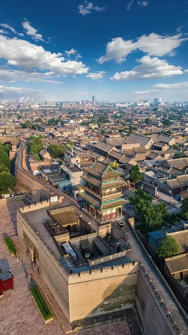 Aerial view of Pingyao Ancient City showing its preserved tall stone city walls with battlements, featuring a prominent multi-tiered traditional Chinese gate tower with green tiled roofs. Inside the city walls, a dense layout of traditional grey-tiled courtyard homes stretches out, with a modern city skyline visible on the horizon under a bright blue sky with white clouds.