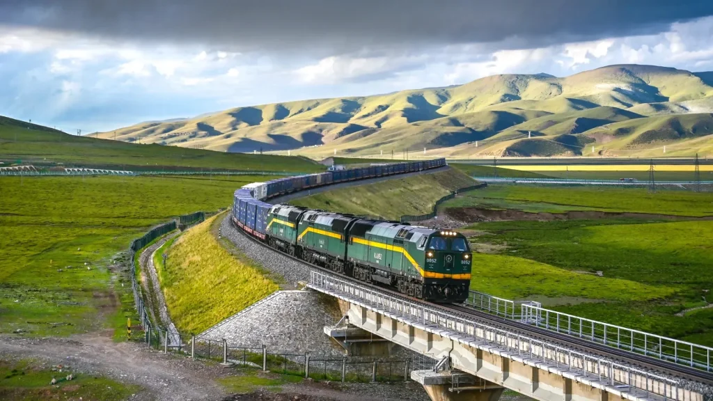 A long green and yellow cargo train with multiple blue containers travels on a curving railway track, crossing a bridge. The tracks wind through a vast, lush green grassland with rolling hills and mountains in the background under a dramatic, partly cloudy sky.