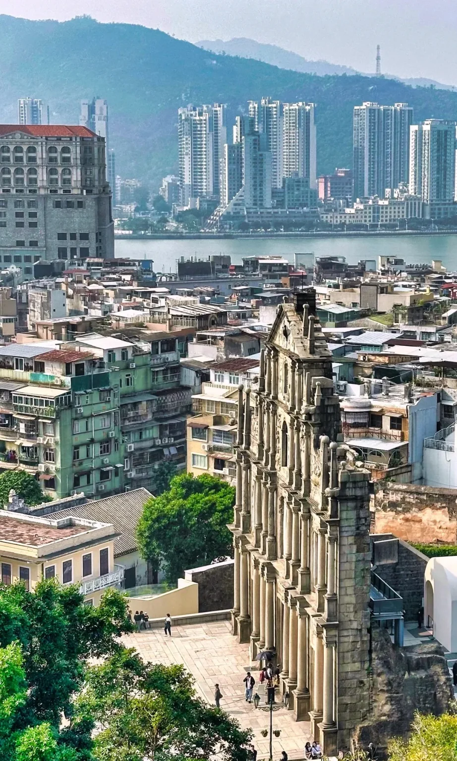 The stone facade of the historic Ruins of St. Paul in Macau, with people walking on the paved plaza below. Behind it, a dense cityscape of traditional and modern buildings stretches towards a body of water and distant hazy mountains.