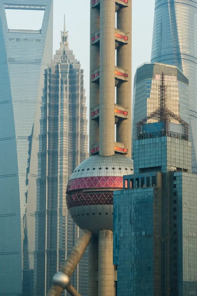 A dense, vertical view of Shanghai's Lujiazui skyline featuring multiple skyscrapers. In the center is the Oriental Pearl TV Tower with its distinctive spheres and concrete support columns. To its left are the tiered, spire-topped Jin Mao Tower and the Shanghai World Financial Center with its trapezoidal opening. Parts of the curved glass Shanghai Tower are visible on the far right.