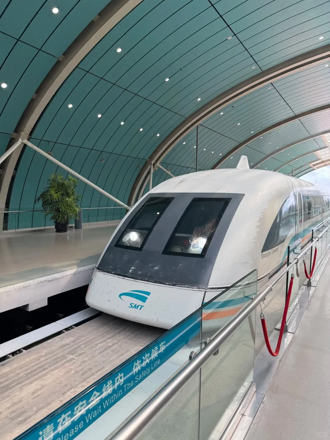 Exterior view of a white Shanghai Maglev high-speed train at a modern station platform. The train features 'SMT' and 'Shanghai Transrapid' branding. On the platform, safety railings, red velvet ropes, and a bilingual sign reading 'Please Wait Within The Safety Line' are visible beneath an arched, light blue ceiling.