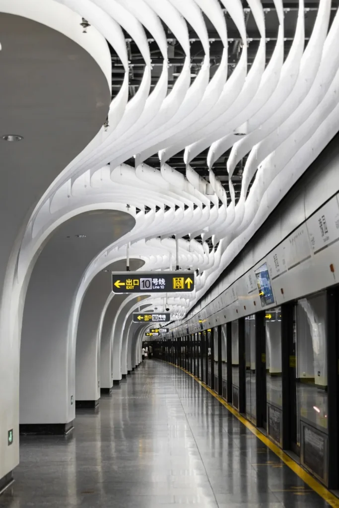 A long, modern subway station platform with a train visible behind platform screen doors on the right. The station features a distinctive ceiling with numerous white, wavy, undulating panels, and curved white structural arches along the left wall. Yellow 'EXIT' and 'Line 10' signs in Chinese and English hang from the ceiling. A bright yellow safety line runs along the edge of the reflective grey platform. A few people are visible in the distance.