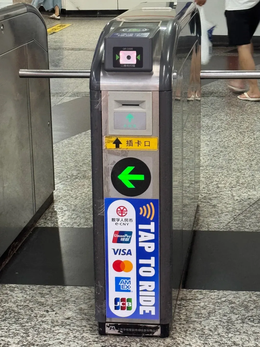 A close-up of a modern gray metro turnstile featuring a QR code scanner on top, a card slot with a green arrow pointing up, and a large green arrow pointing left below it. A vertical sticker on the side lists various payment logos including e-CNY, UnionPay, Visa, Mastercard, American Express, and JCB, next to the text 'TAP TO RIDE' and a contactless payment symbol. The turnstile has reflective metal surfaces, and the legs of a person are partially visible in the background.