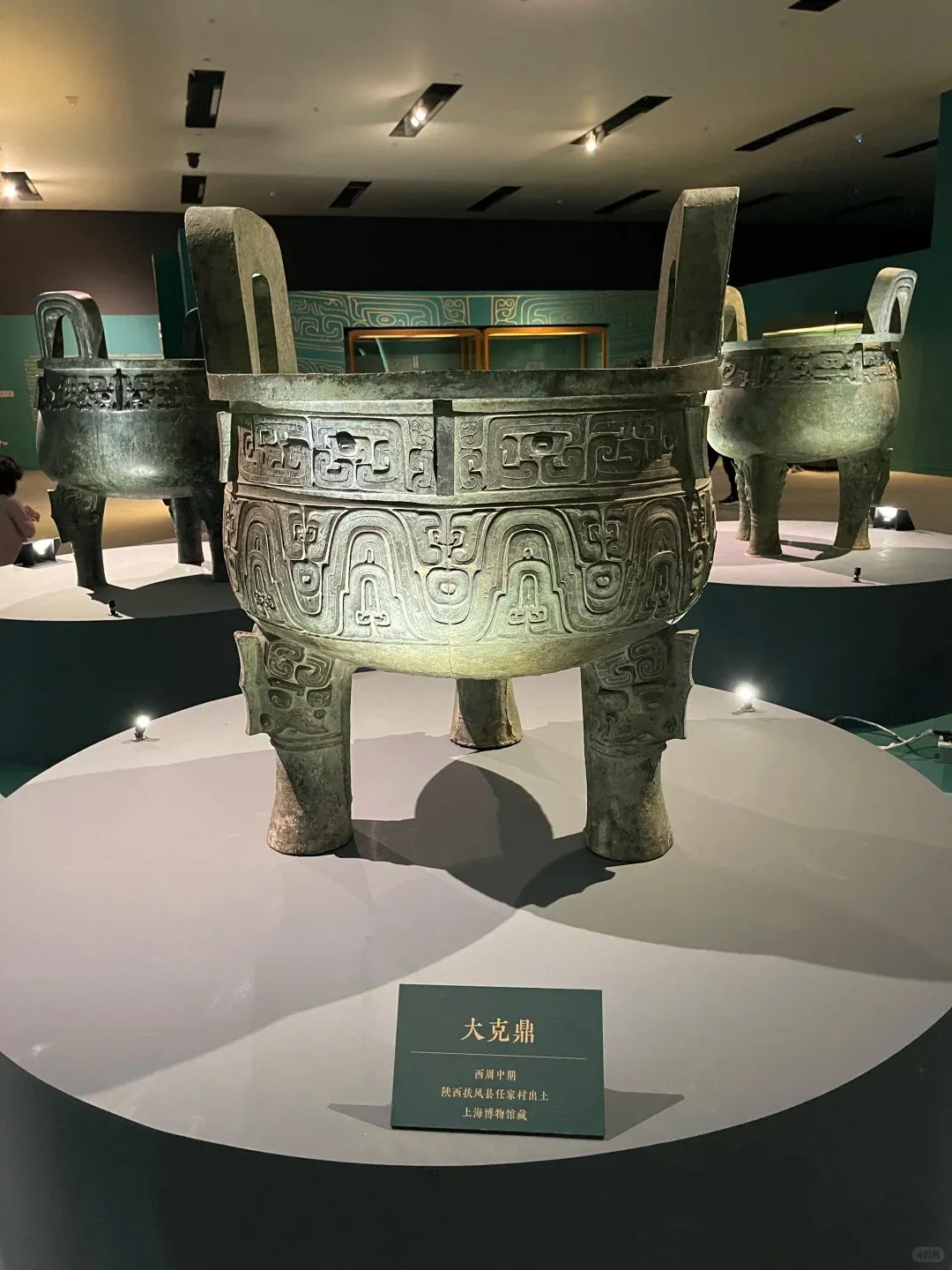A large ancient Chinese bronze ding vessel with three legs, two upright handles, and intricate swirling patterns, displayed on a circular gray pedestal in a dimly lit museum gallery. Two smaller bronze vessels are visible in the background, and a display sign in front identifies the main artifact as the Da Ke Ding from the Mid-Western Zhou Dynasty.