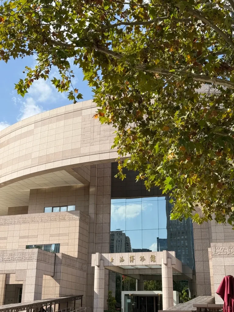 Exterior view of the Shanghai Museum building, featuring its light-colored stone tiled facade with a curved upper section and a prominent glass entrance reflecting sky and nearby buildings. A large tree with green and some brown leaves partially frames the top of the museum against a blue sky.