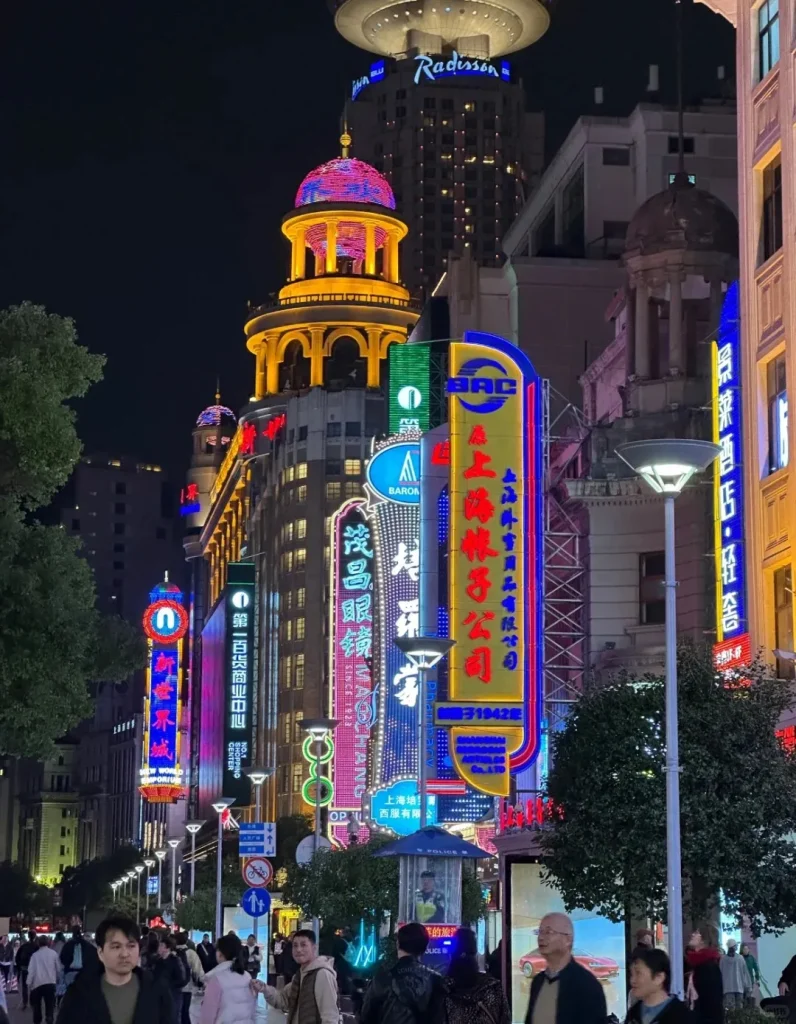 A vibrant night scene on Nanjing Road in Shanghai, China, with numerous brightly lit neon signs in Chinese characters adorning buildings, including ornate historical architecture and the modern Radisson Blu hotel with a distinctive circular rooftop structure. Pedestrians fill the bustling street.