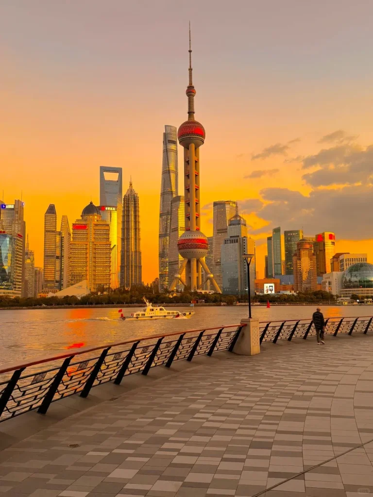 Panoramic view of the Shanghai Pudong skyline at sunset, featuring the red-and-white Oriental Pearl TV Tower, the Shanghai Tower, and other modern skyscrapers. The Huangpu River flows in the foreground with a white boat, and a person walks on a tiled promenade with a decorative black and red railing.