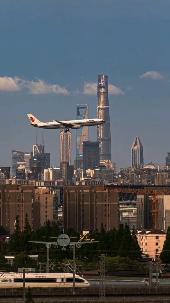 A white Air China passenger plane with a red and blue phoenix logo flies across the modern Shanghai skyline, featuring the Shanghai Tower and Shanghai World Financial Center under a blue sky. In the foreground, a high-speed train blurs past on elevated tracks, with residential buildings and green trees below.