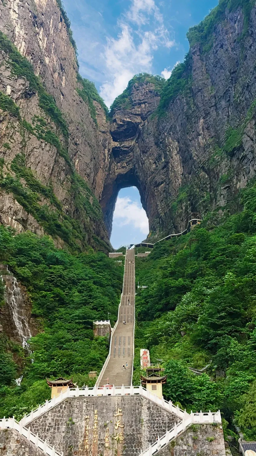 A very long and steep stone staircase, known as the Stairway to Heaven, leads up a lush green mountain to a large natural rock arch called Tianmen Cave or Heaven's Gate. Tall, rocky cliffs covered in dense green foliage flank both sides. A small waterfall cascades down the cliff on the left. People are visible ascending the stairs, and an escalator system runs parallel to the right side of the staircase. A traditional building with an ornate roof stands at the bottom of the steps.