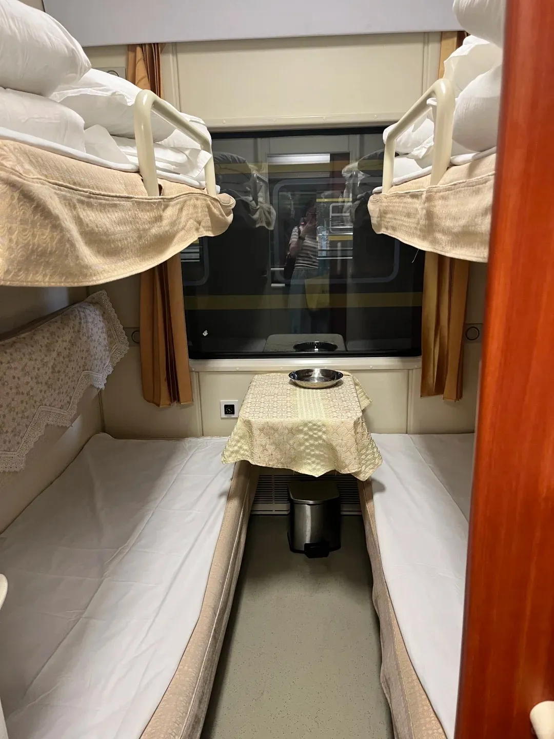 Interior of a soft sleeper cabin on a train, featuring two lower bunks with white sheets and two upper bunks with bedding stacked. A small table with a patterned tablecloth stands between the lower bunks, topped with a silver bowl, and a small pedal bin sits underneath. A large window with drawn curtains reflects the photographer.