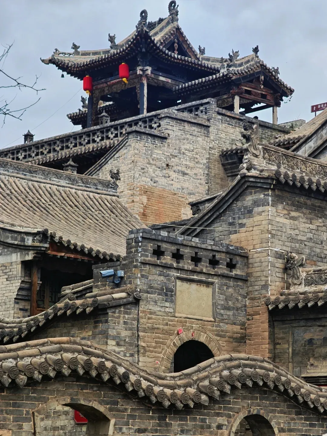 A complex of ancient Chinese architecture featuring dark grey brick walls and multiple tiled roofs. The tallest structure is a multi-tiered pagoda with dark eaves and two bright red lanterns hanging from its upper level. Intricate carvings of mythical creatures adorn the roof ridges of several buildings, which include arched doorways and decorative wall sections.