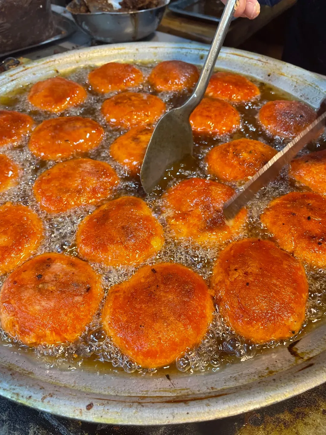 Numerous round, orange-red persimmon cakes deep-frying in a large metal pan filled with bubbling oil. A metal spatula and tongs are visible, turning the cakes.