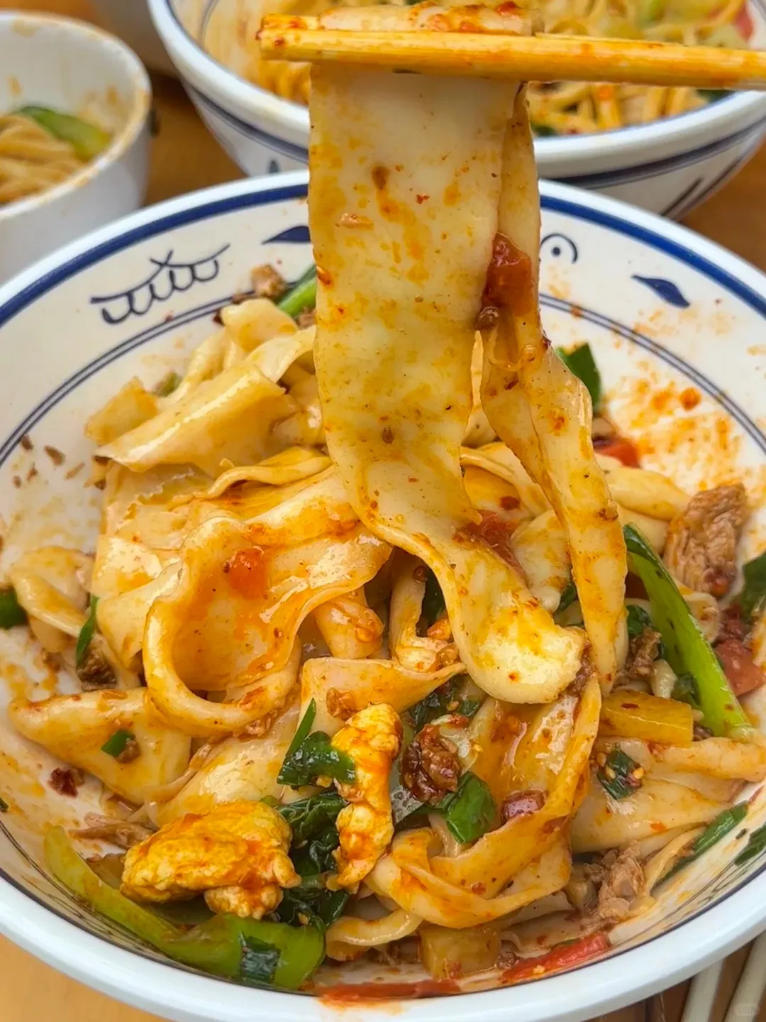 Chopsticks lifting a wide, hand-pulled Biang Biang noodle from a white bowl. The noodles are tossed in a vibrant red chili oil sauce with green vegetables, scrambled egg, and bits of minced meat visible.