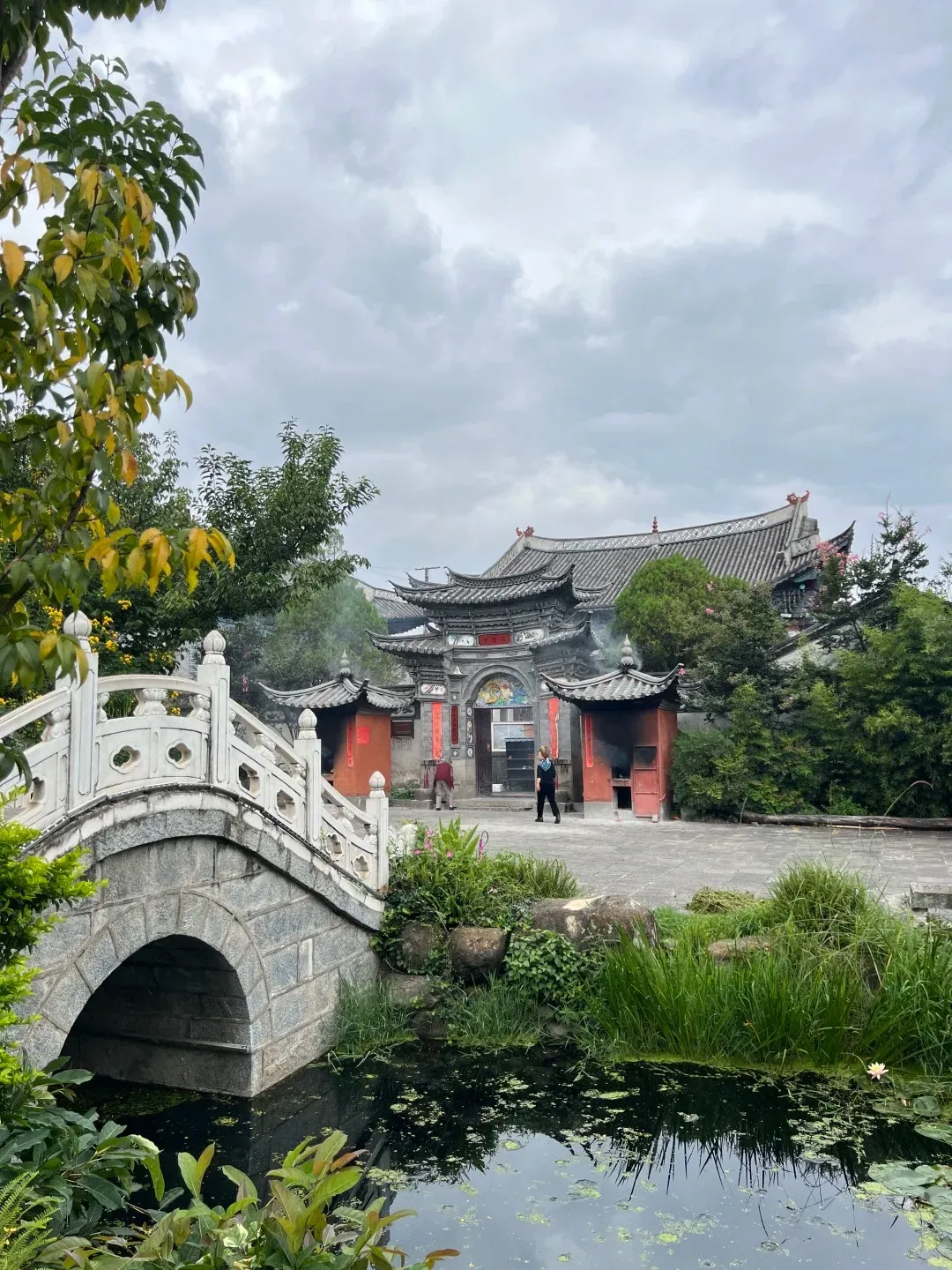 A traditional stone arch bridge with white railings crosses a dark pond filled with green lily pads and water plants. In the background, a traditional Chinese temple complex features multiple dark tiled roofs, red architectural details, and smoke rising from two outdoor offering structures. Several people are visible near the temple entrance under an overcast sky.